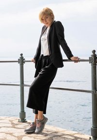 Schwarzes Blazer über einem weißen Grafik-T-Shirt, kombiniert mit schwarzen Hosen und grauen Sneakers mit rosa Schnürsenkeln. Steht an einem Geländer am Wasser.