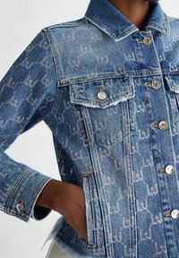 Jeansjacka i ljusblå färg, silverfärgat logomönster, klassisk krage, tryckknapps-stängning och framfickor med fransade sömmar.