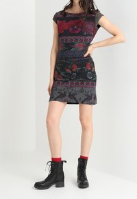 Robe ajustée à manches courtes avec un motif floral foncé, fond noir et accents colorés. Associée à des bottes noires à lacets et des chaussettes rouges.
