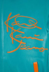 Tyrkysová kožená bunda s reliéfním oranžovým podpisem. Hladký povrch s kontrastním šitím a výrazným nášivkou.