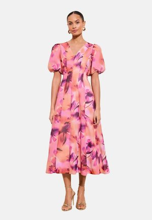 Femeie purtând o rochie midi roz cu imprimeu floral, mâneci scurte bufante și decolteu în V, asortată cu sandale aurii cu barete și toc înalt.