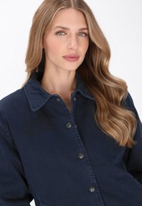 Sötétkék farmerdzseki, galléros nyakkal, hat fém gombbal, és finom hímzett szöveggel a bal oldalon. Simaságáért kényelmes tapintású.