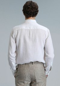 Chemise en lin de couleur claire avec des manches longues et un col classique, présentant une texture subtile. Associée à un pantalon beige rayé.