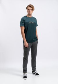 Teal katoenen t-shirt met een grafische print op de borst, gecombineerd met grijze jogger pants, zwarte sneakers met witte accenten en minimale details.