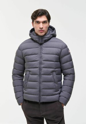 Mann trägt eine graue Steppjacke mit Kapuze, Hände in den Taschen, steht vor einem schlichten hellen Hintergrund.