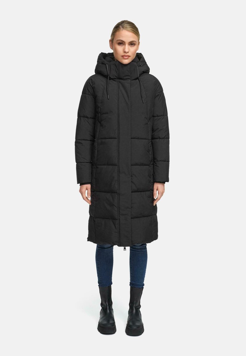 Sort lang puffersigte med hætte, der har store quiltede sektioner, en for-lynlås og sidelommer, parret med mørke jeans og ankelstøvler.