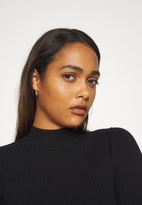 Black ribbed high-neck top with long sleeves, featuring a fitted design and subtle texture. Model wears small hoop earrings.