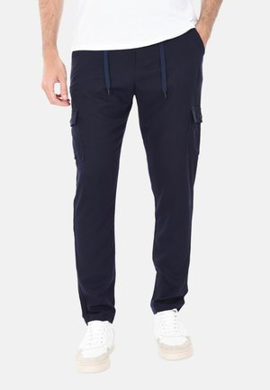 Uomo che indossa pantaloni cargo blu navy con tasche laterali, una t-shirt bianca e scarpe da ginnastica bianche, in piedi con una mano in tasca.