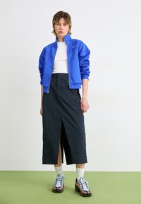 Blå zip-jacka, vit åtsittande skjorta, mörk kjol med framklipp och svart-orange sneakers med blåa accenter. Grön golvbakgrund.