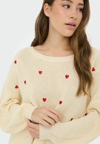 Jeune femme aux longs cheveux portant un pull en maille crème décoré de petits cœurs rouges, regardant sur le côté, tenant la manche.