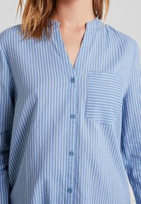 Camicia azzurro chiaro con bottoni, caratterizzata da strisce bianche verticali, colletto a V e una tasca sul lato sinistro con strisce orizzontali.