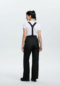 Kobieta z ciemnym kucykiem, ubrana w białą koszulę z krótkim rękawem i czarne spodnie z szelkami, zwrócona plecami na jednolitym jasnym tle.