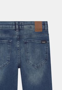 Dark blue denim jeans feature two back pockets with a subtle wash effect. A brown leather tag embossed with "CARS" is at the waistband.