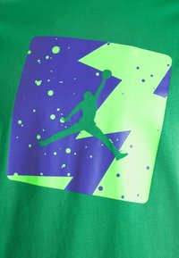 Zielony T-shirt z fioletową i neonowozieloną grafiką skaczącej postaci, otoczoną wzorami rozbryzgów i kanciastymi kształtami.