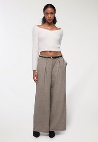 Una donna è in piedi indossando un maglione corto bianco fuzzy senza spalline, pantaloni beige a vita alta e gamba larga con cintura nera e scarpe nere con punta.