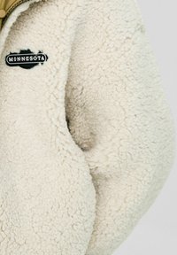 Nærbillede af en fløjlsk jacket i cremefarve med et sort og hvidt "MINNESOTA" patch nær lynlåsen på venstre side.