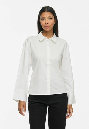 Femme aux longs cheveux foncés portant une chemise blanche à col et manches longues et un pantalon noir, debout devant un fond blanc uni.