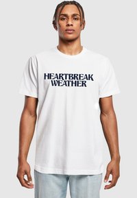Hvit bomullst-skjorte med mørk marineblå tekst "HEARTBREAK WEATHER" i en fet, moderne skrifttype. Korte ermer og rund hals.