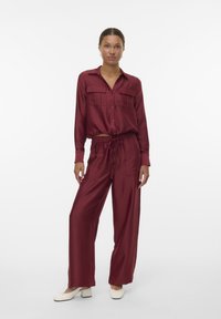 Burgundy, long-sleeve shirt with two chest pockets and drawstring, wide-leg pants. Smooth texture; model wears cream block-heeled shoes.