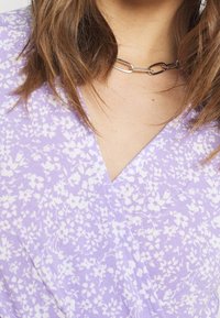 Tissu violet à motif floral avec un décolleté en V, présentant de petites fleurs blanches. Collier en chaîne argentée visible au niveau du col.