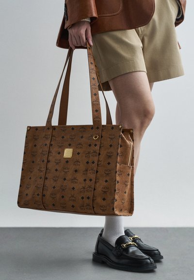 Osoba držící hnědou tašku s potiskem a zlatou destičkou, v béžových šortkách, hnědé bundě, bílých ponožkách a černých mokasínech se zlatým detailem.