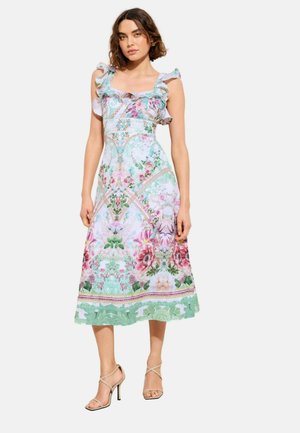 Ženska stoji v brezrokavni, nabrani midi obleki s pastelno cvetličnimi in dekorativnimi vzorci, v kombinaciji z bež sandali s trakovi in visokimi petami.