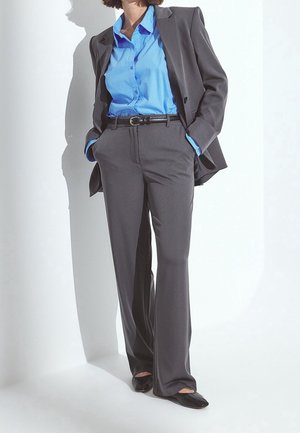 Personne portant un costume gris foncé avec un pantalon assorti, une chemise bleu clair boutonnée, une ceinture noire et des chaussures noires à bout pointu, les mains dans les poches.