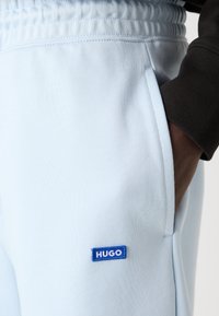 Šviesiai mėlyni sportiniai džinsai su raišteliu juosmens srityje, šonine kišene ir mėlyna etiketė "HUGO" priekyje. Lygus paviršius.