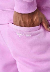 Roze joggers van stof met een gladde textuur, voorzien van een zijzak en geborduurd "Project x Paris" logo in het wit nabij de rand van de zak.