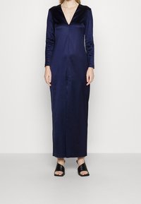 Long navy satin dress with a deep V-neck and long sleeves. Features a smooth texture and a straight silhouette. Paired with black open-toe sandals.