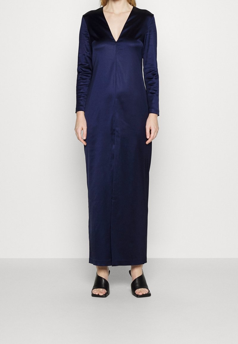 Long navy satin dress with a deep V-neck and long sleeves. Features a smooth texture and a straight silhouette. Paired with black open-toe sandals.