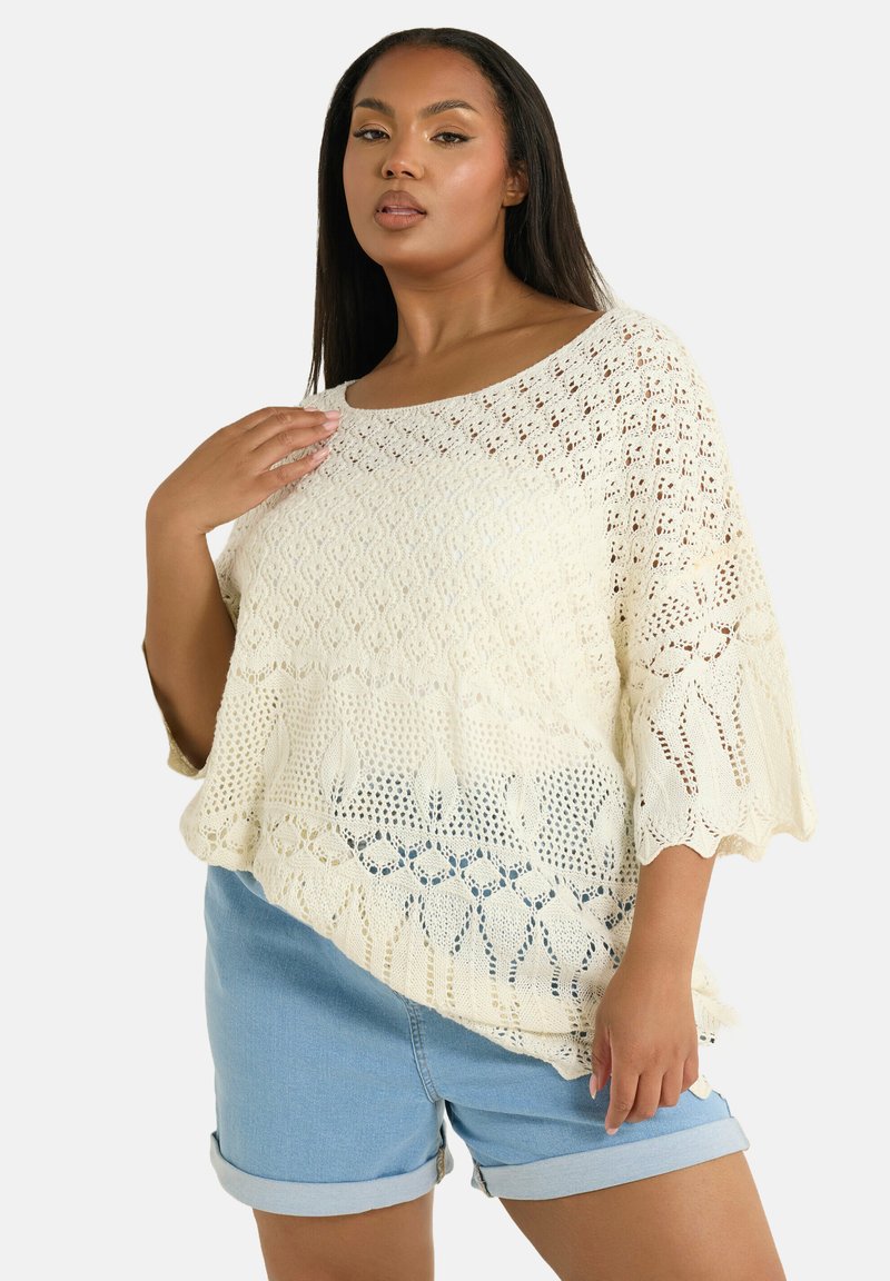 Maglione crema con motivo a rete aperta, scollatura ampia e maniche a onda, abbinato a pantaloni corti azzurri con orlo arrotolato.