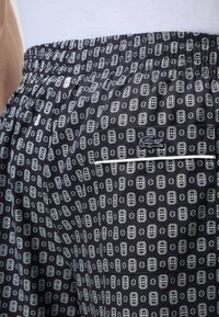Gros plan sur un short noir avec motif géométrique blanc, taille élastique, poche latérale et petit logo crocodile brodé.