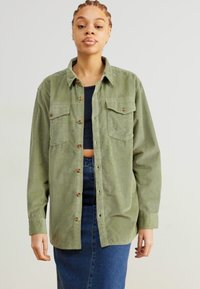 Grøn kord-urot skjorte med knaplukning foran, to brystlommer og en afslappet pasform, parret med en blå denimnederdel.