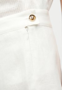 Άσπροι υφασμάτινοι παντελόνια με πλευρικό φερμουάρ και πόρπη με χρυσό κουμπί, φορεμένα με ανοιχτόχρωμο ριμπ τοπ.