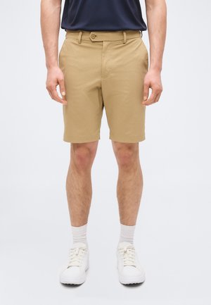 Bărbat purtând pantaloni scurți bej până la genunchi, șosete albe, adidași albi și o cămașă bleumarin, stând în fața unui fundal simplu.