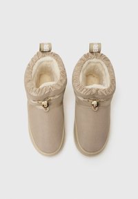 Beige, kényelmes slip-on csizmák, textúrált anyagból készült, bolyhos béléssel és elasztikus tetejével. Díszes arany rögzítőelemmel a beállításhoz.