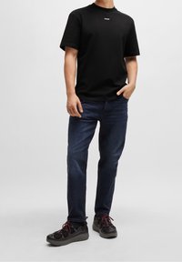 Zwarte t-shirt met korte mouwen en een wit logo op de borst, gecombineerd met donkerblauwe jeans en zwarte schoenen met rode veters en een gestructureerd design.