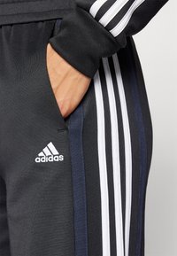 Crne atletske hlače s bijelim i mornarskoplavim prugama, s bočnom džepom i bijelim Adidas logotipom na donjoj lijevoj nozi.