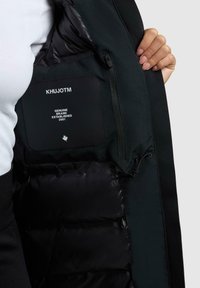 Schwarze Jacke mit einer strukturierten Außenschicht und glattem Innenfutter. Verfügt über ein Etikett mit "KHUJOTM", einer Reißverschlusstasche und einem Kordelzug-Detail.