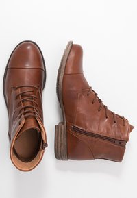 Botas de cuero marrón hasta el tobillo con punta redonda, cordones en la parte delantera y cremallera lateral. El tacón apilado y la suela texturizada ofrecen mayor agarre.