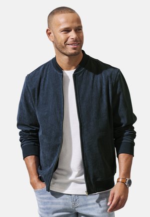 Lächelnder Mann mit dunkelblauer Zip-up-Jacke, weißem T-Shirt, hellen Jeans und brauner Uhr, die Hände entspannt an den Seiten.