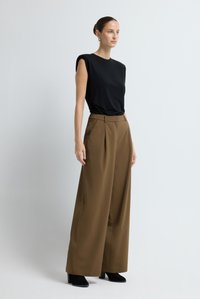 Zwarte top met korte mouwen en bruine, hooggetailleerde broek met brede pijpen. Gevlochten stof met strakke lijnen. Zwarte laarzen met hak op een neutrale achtergrond.