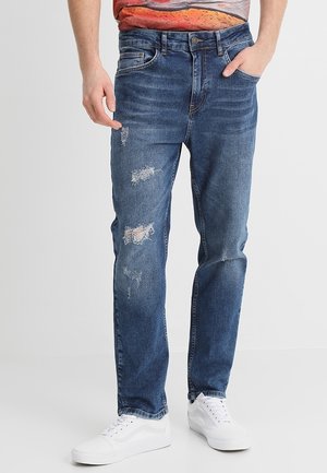 Homme portant un jean bleu déchiré et des baskets blanches, une main dans la poche, sur un fond uni.