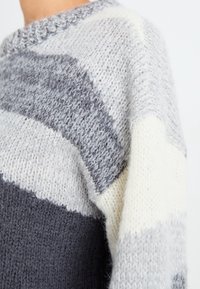 Pull tricoté avec des rayures grises, blanc cassé et gris foncé. Tissu texturé avec un col rond et des détails de couture visibles.
