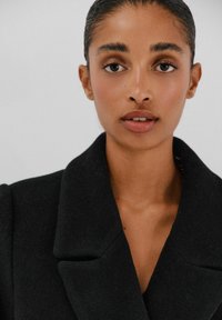 Musta takki, jossa on leveä kaulus ja teksturoitu kangas. Lähikuva kasvosta, jossa on sileä iho, neutraalit huulet ja tummat kulmakarvat.