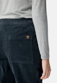 Marineblaue Cordhosen mit strukturierter Oberfläche, ausgestattet mit einer aufgesetzten Gesäßtasche und einem kleinen, hellbraunen Etikett. Kombiniert mit einem hellgrauen Langarmshirt.