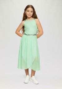 Mintgroene jurk met mouwloos ontwerp, glinsterende tailleband en een vloeiende, gelaagde rok. Gecombineerd met witte sneakers voor een casual uitstraling.