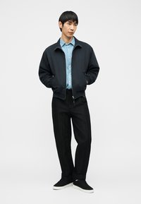 Νεαρός άνδρας στέκεται με τα χέρια στις τσέπες, φοράει μαύρο σακάκι, ανοιχτό μπλε πουκάμισο με κουμπιά, μαύρο παντελόνι και μαύρα αθλητικά παπούτσια με λευκές σόλες.