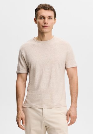 Ung mand med kort brunt hår iført en beige T-shirt med korte ærmer og lyse bukser, står mod en ensfarvet baggrund.